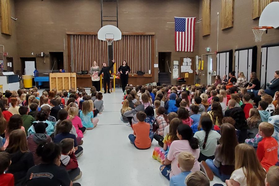 Three performers entertain children in a school gym with a brick wall backdrop.