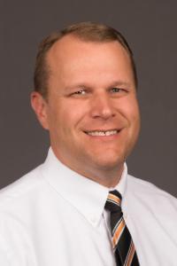 A medium skin toned man with balding brown hair. He is wearing a white shirt and black tie