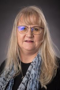 A light skin toned woman with glasses and blond hair. She is wearing a black shirt and a blue scarf