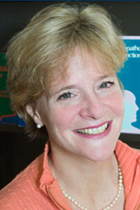 A light skin toned woman with short blond hair wearing a coral shirt. She is smiling