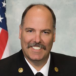 A medium skin toned man with balding brown hair and a mustache. He is wearing a button down, tie and suit jacket. He is smiling