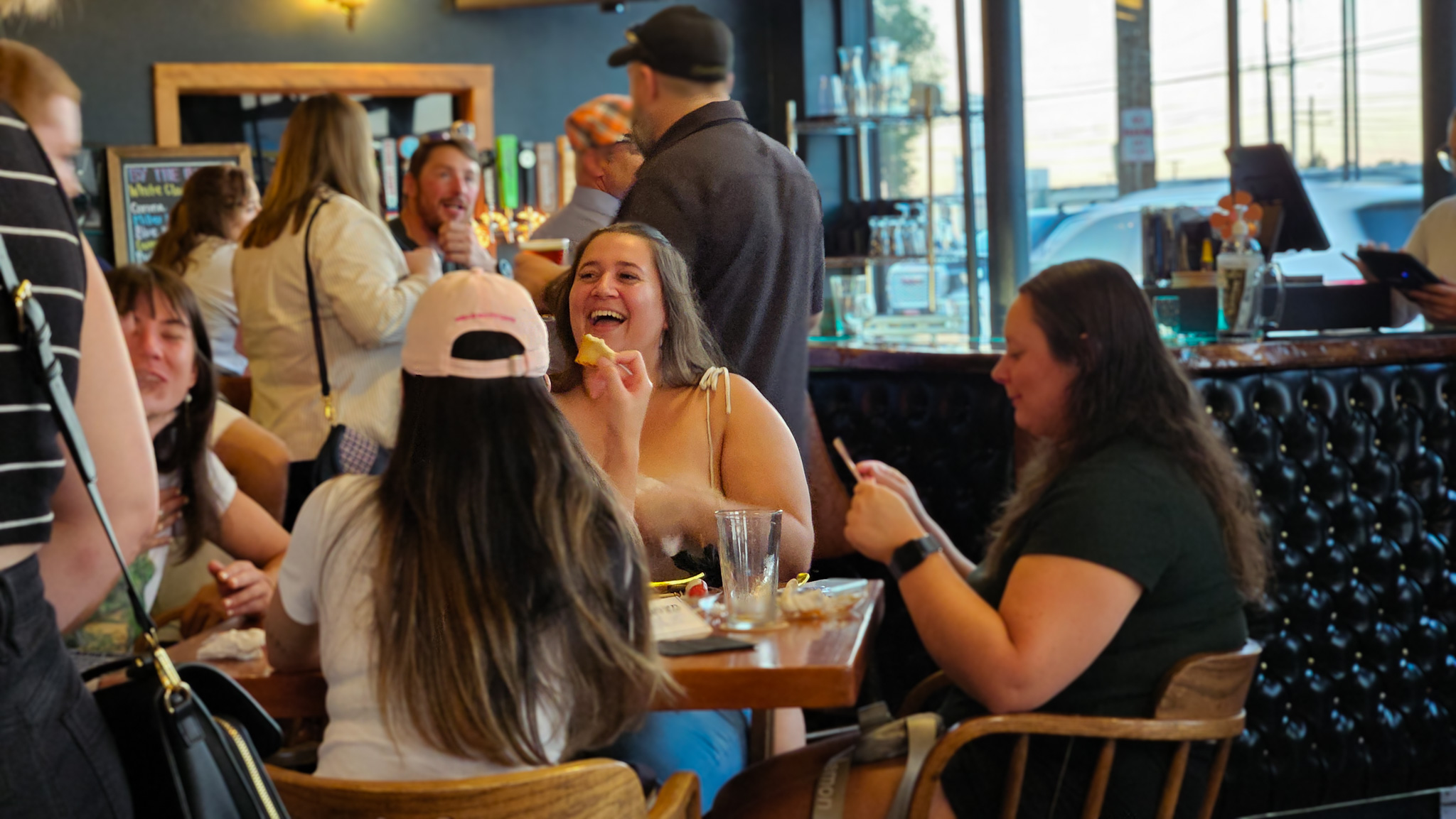4 people laughing and sitting at a table in a bar, talking to someone standing near the table