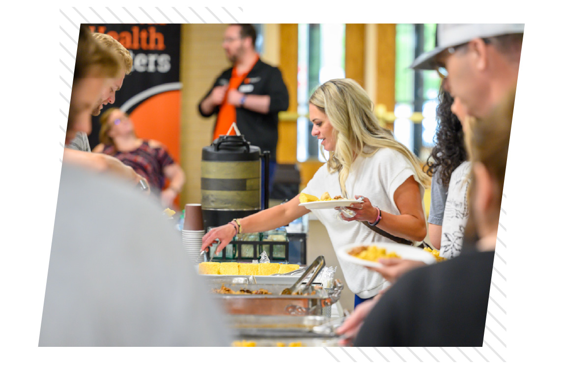 A group of people serving themselves food from a catered buffet table inside an indoor event space. One person holds a plate while selecting food, with beverage dispensers and other attendees visible in the background.