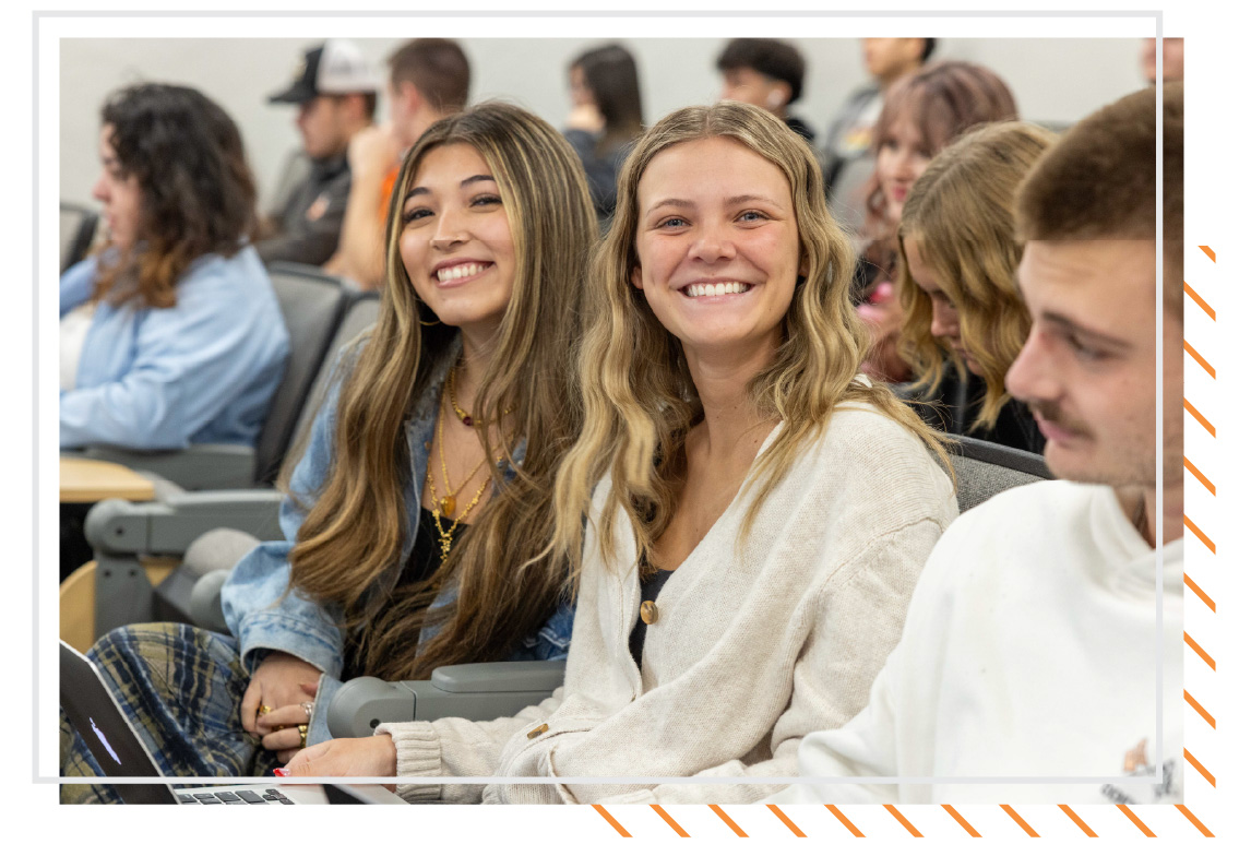 Two students sitting in a classroom at Idaho State University Idaho Falls, smiling toward the camera, with other students seated in the background.