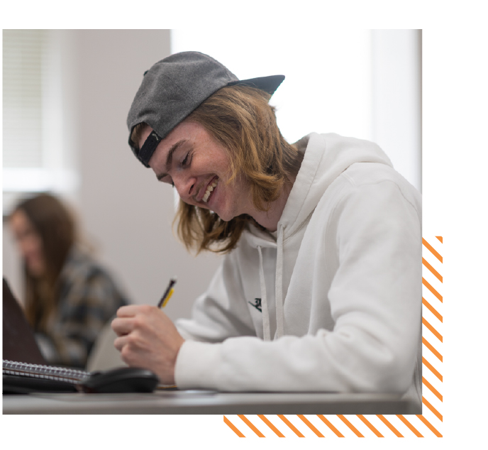 College student smiling while taking notes at a desk with a computer in a classroom, representing hands-on learning and academic support in ISU’s associate degree programs.