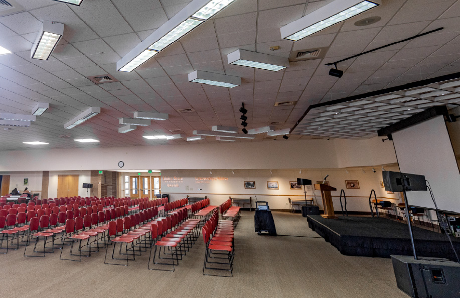 Wide interior view of a large auditorium set up with rows of red chairs facing a raised stage with a podium, projection screen, and audio equipment; acoustic ceiling panels, wall signage, and rear entrance doors identify the Idaho State University Idaho Falls venue, suitable for lectures and community events