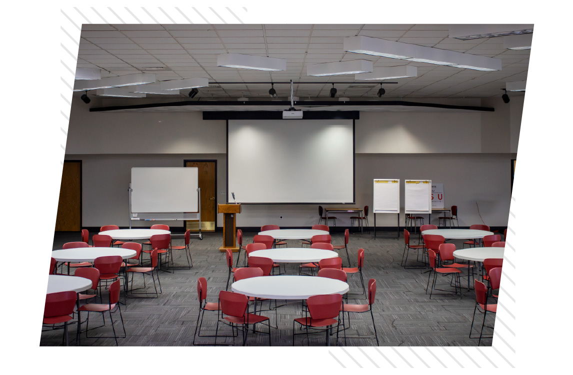 Spacious multipurpose room set up with round tables and red chairs, facing a large projection screen, podium, whiteboard, and flip charts at the front.