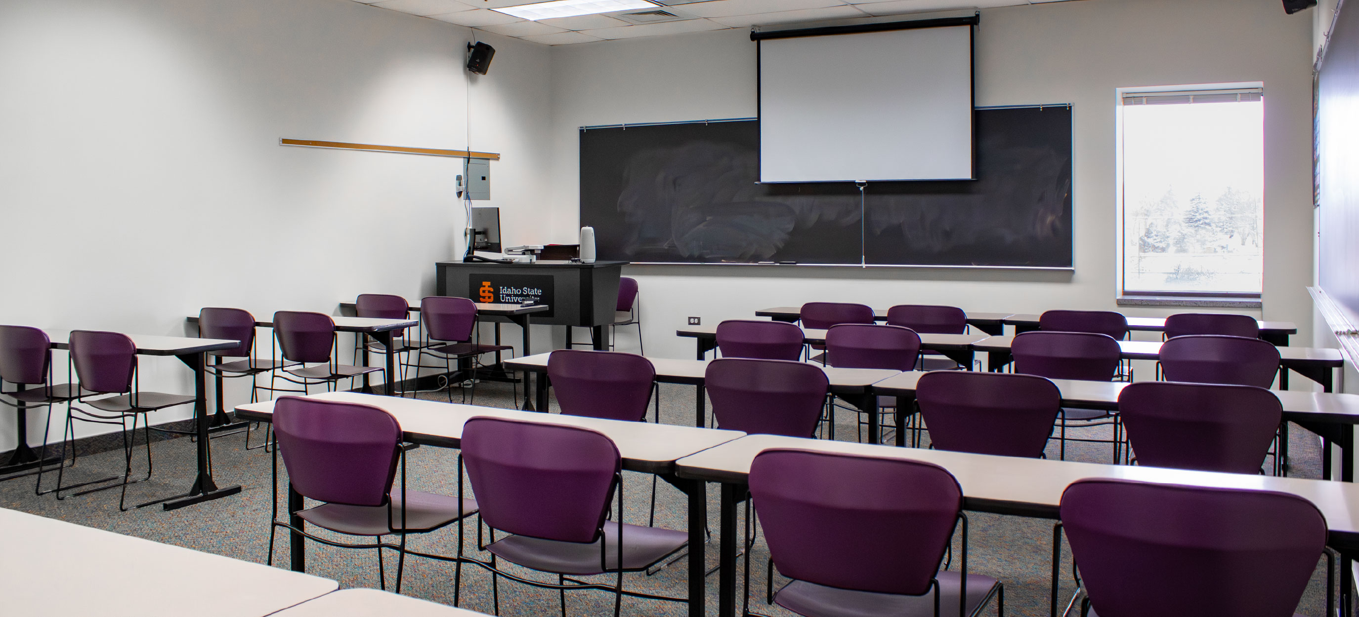 CHE classroom with rows of tables and purple chairs facing a teaching station, chalkboards, and a pull-down projection screen.