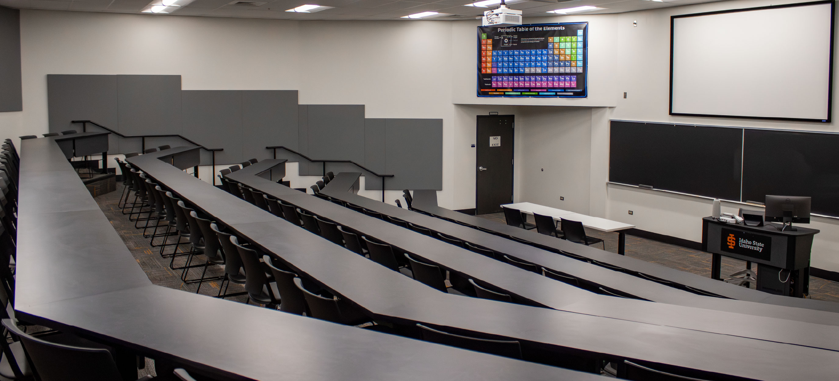 Wide view of the CHE Large Tiered Classroom at ISU Idaho Falls, showing curved rows of fixed tables and chairs facing blackboards, a projection screen, instructor station, and periodic table display.