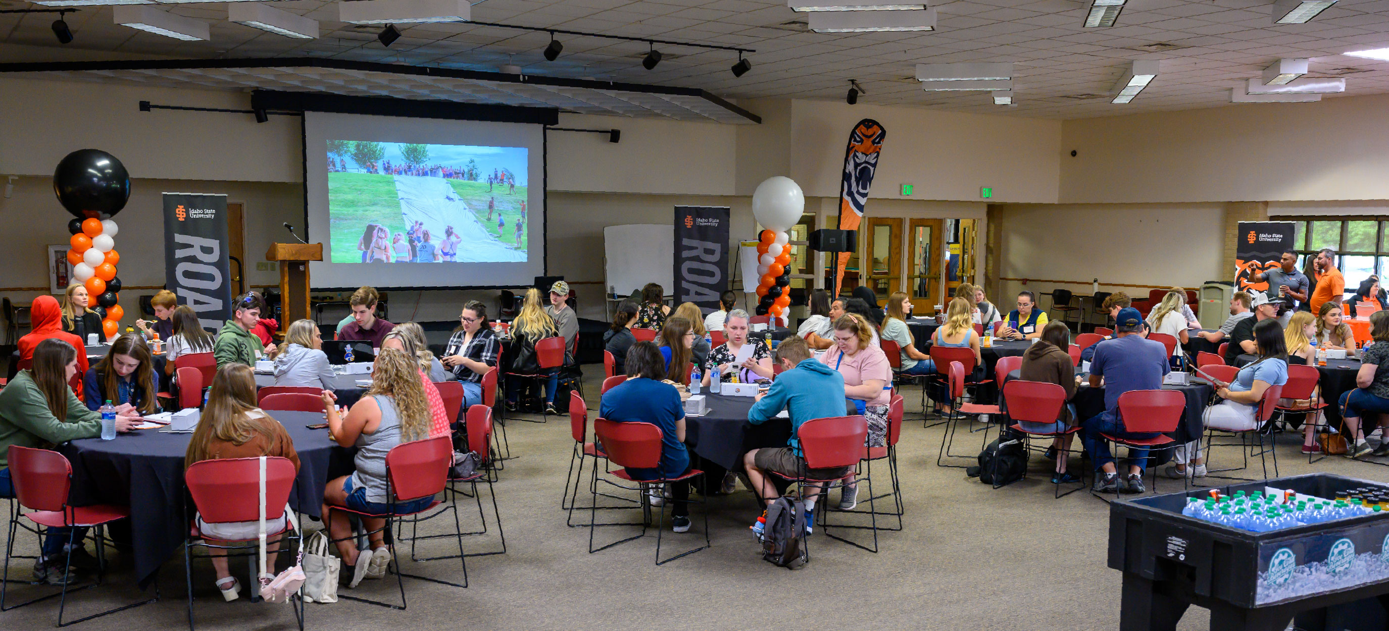 Large event space set up banquet-style with round tables and red chairs, filled with attendees seated and talking, with Idaho State University banners, balloon decorations, and a presentation displayed on a projection screen at the front.