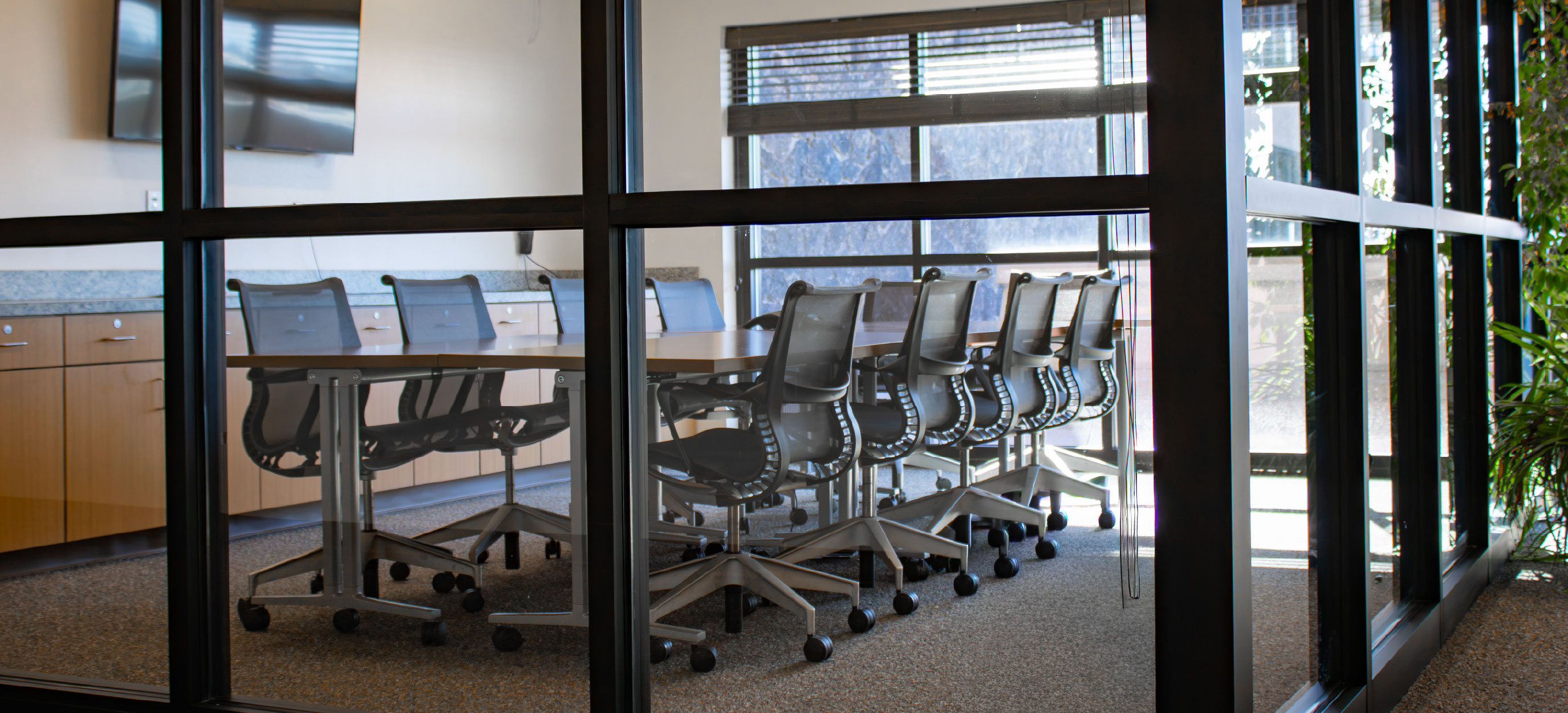 Conference room viewed through glass walls, featuring a long meeting table, black rolling chairs, a wall-mounted display, built-in cabinetry, and large windows.