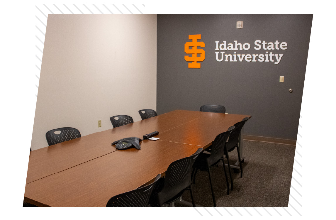 Conference room with a long wooden table, black chairs, a conference phone, and an Idaho State University logo displayed on the dark gray wall.
