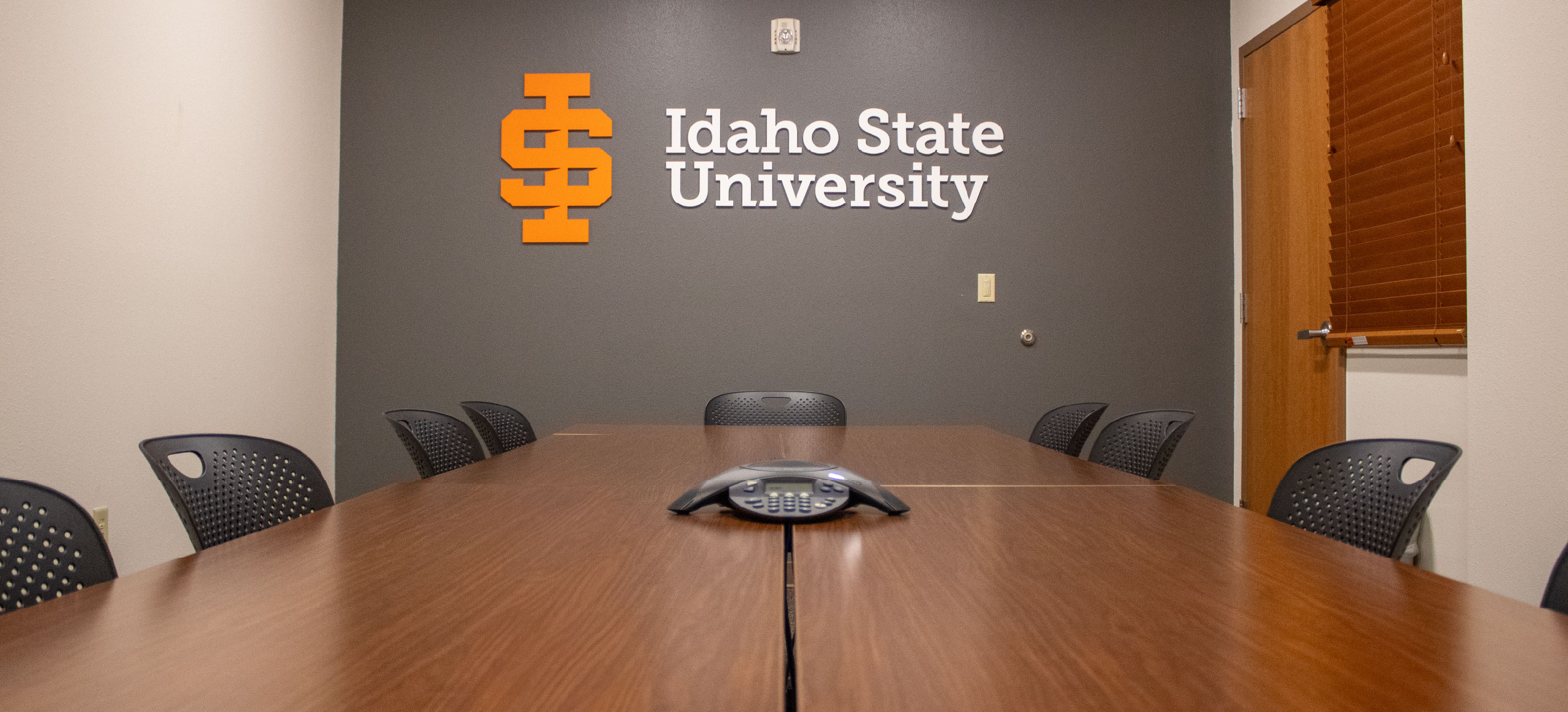 TAB 279 conference room with a large wooden table, black chairs, a conference phone, and the Idaho State University logo displayed on the back wall.