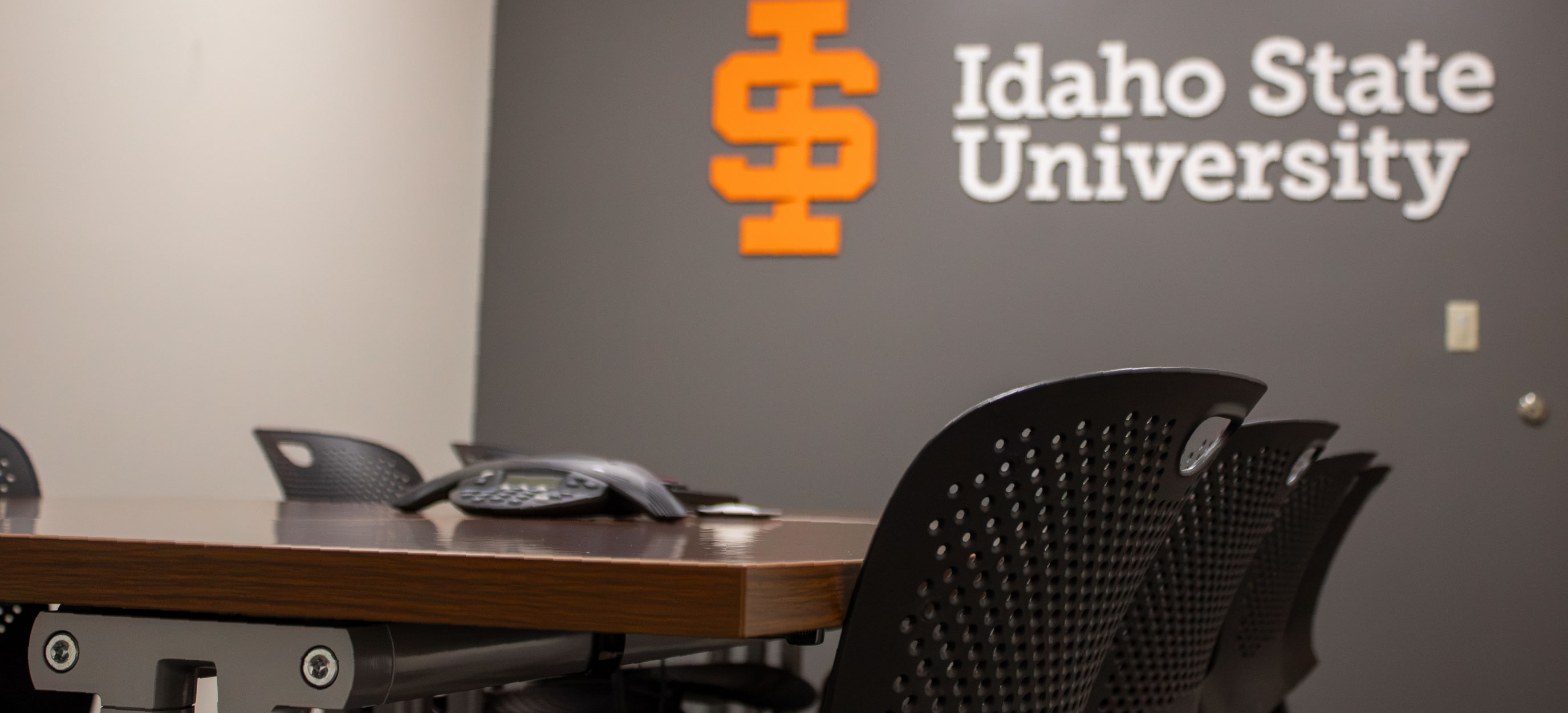 Close-up of the TAB 279 conference room showing a wooden conference table, black chairs, a conference phone, and the Idaho State University logo on the wall.