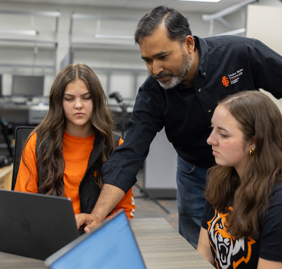 An Idaho State University instructor leans over a desk to assist two female students with a laptop in a lab setting. The instructor wears a black shirt featuring, while the students wear ISU-themed apparel, including an orange shirt and a black tee.