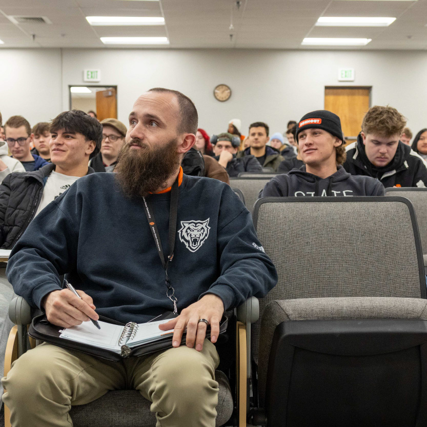 Students seated in a classroom lecture, with one student in the foreground taking notes while others listen attentively.