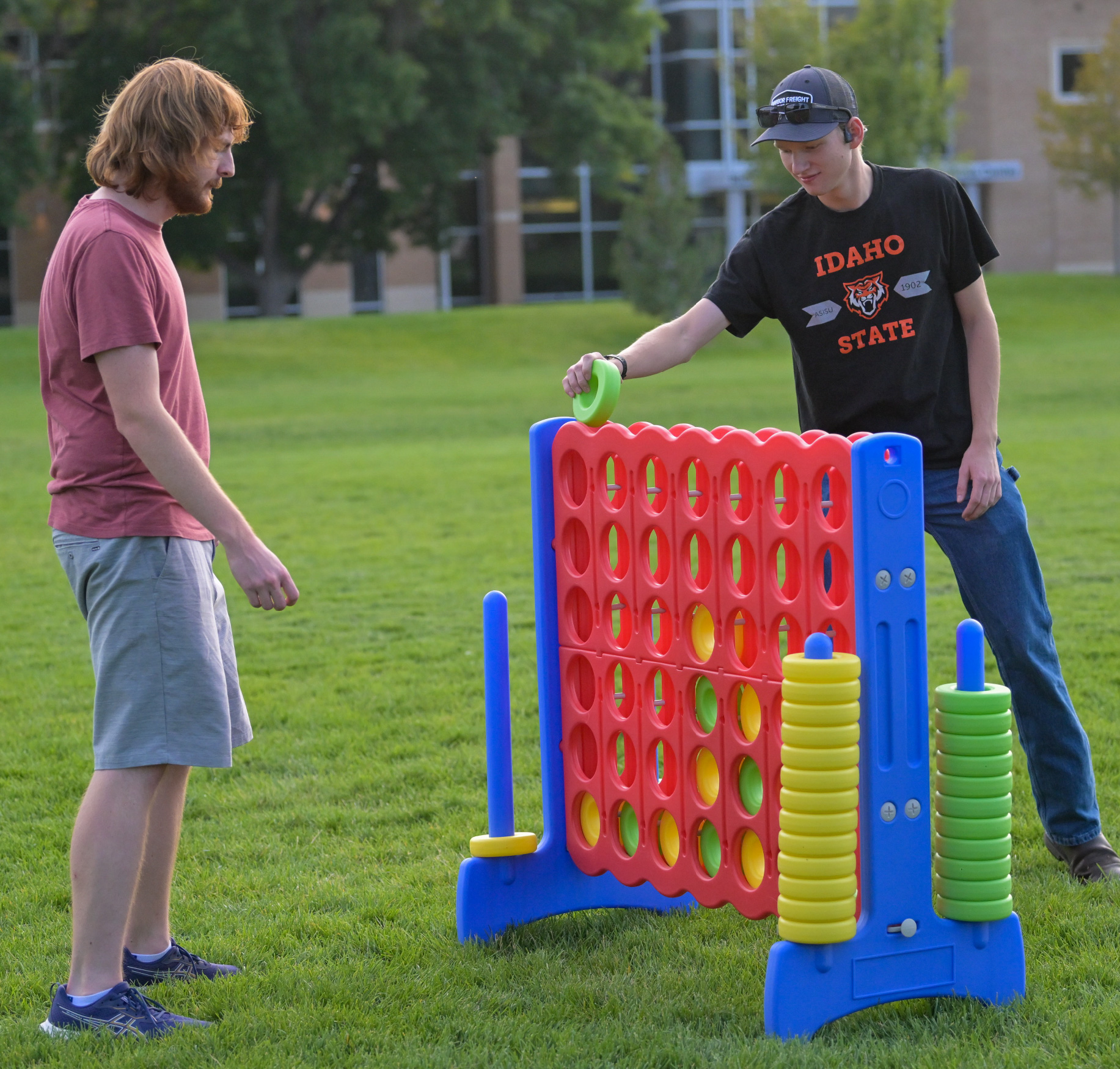 Two college students play a large outdoor Connect Four game on a grassy campus lawn. One student places a green game piece into the board while the other watches, with campus buildings and trees in the background, creating a fun and relaxed campus atmosphere.
