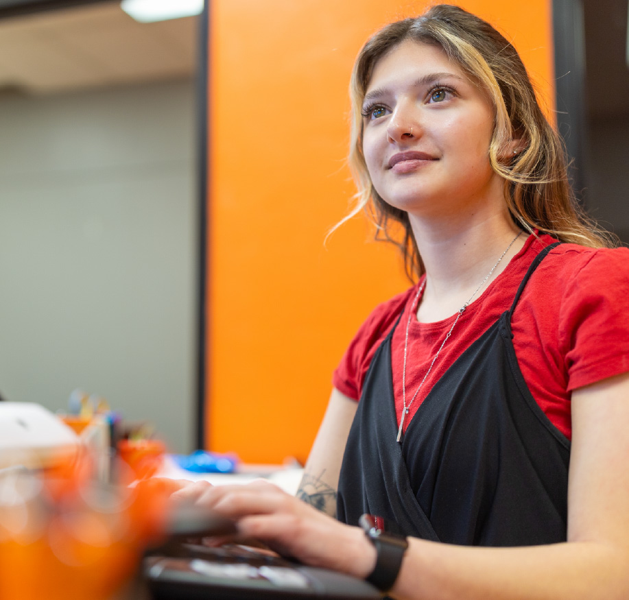 A college student sits at a desk in a classroom or computer lab, looking attentive and focused. An orange accent wall and desk items are visible in the background, creating a modern and engaged learning environment.