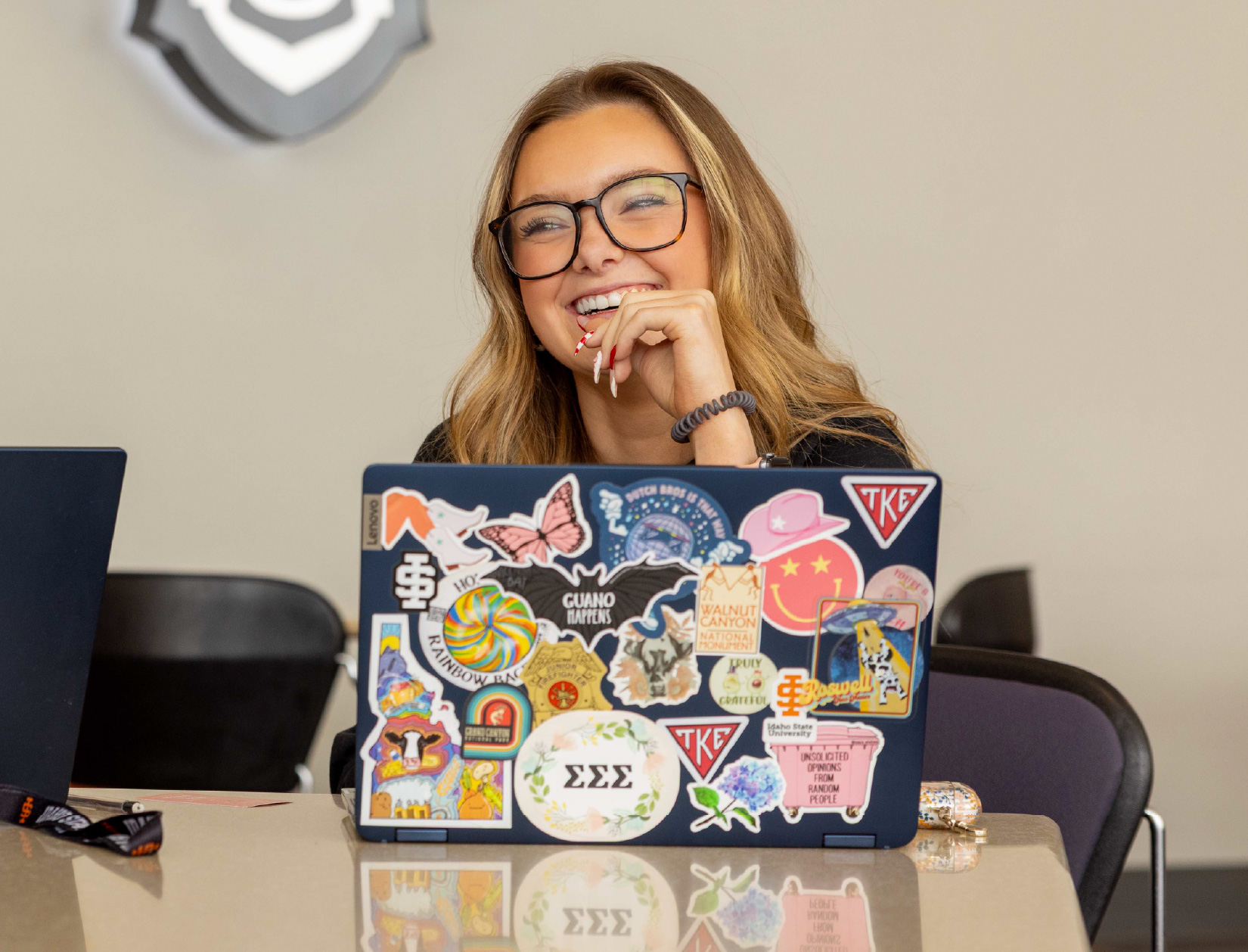 Smiling college student wearing glasses sits at a table with a sticker-covered laptop, laughing while working in a classroom or study space.
