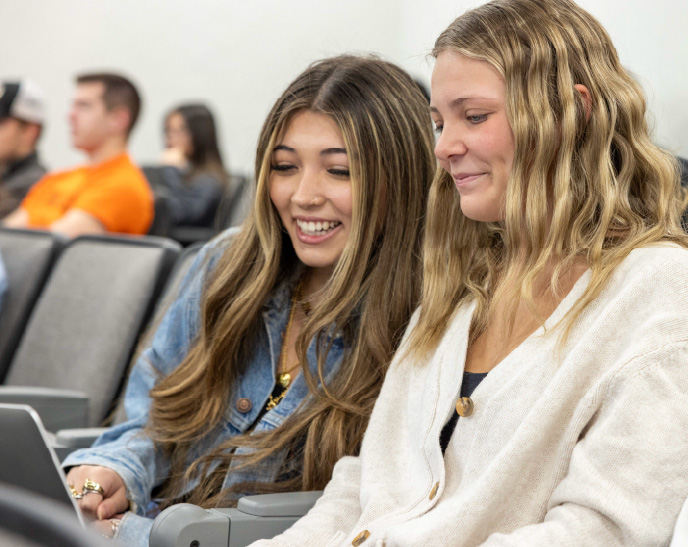 Two students sit together in a classroom, smiling as they look at a laptop during class. Other students are seated in the blurred background, creating a collaborative and welcoming learning environment.