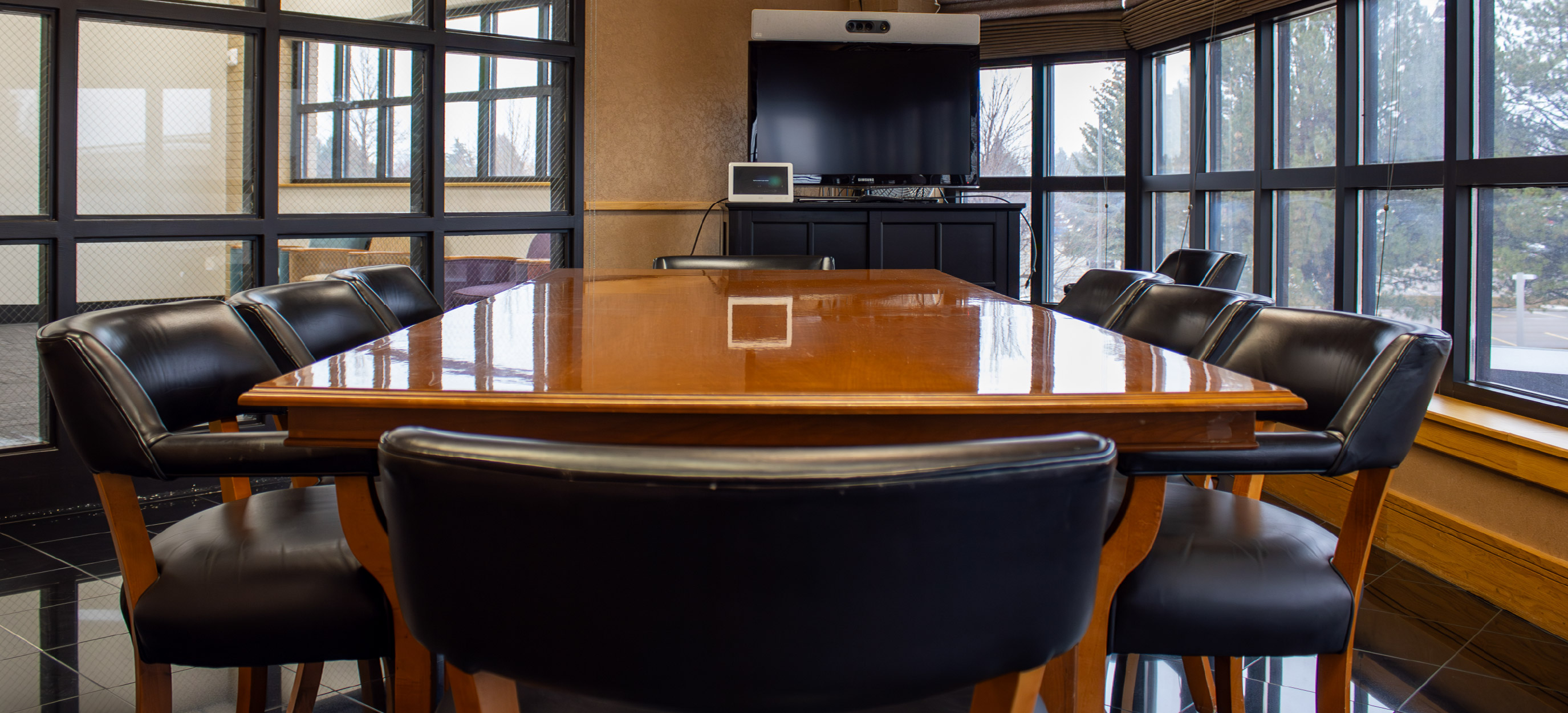Conference room with a large polished wooden table, black leather chairs, floor-to-ceiling windows, and a wall-mounted display for presentations.