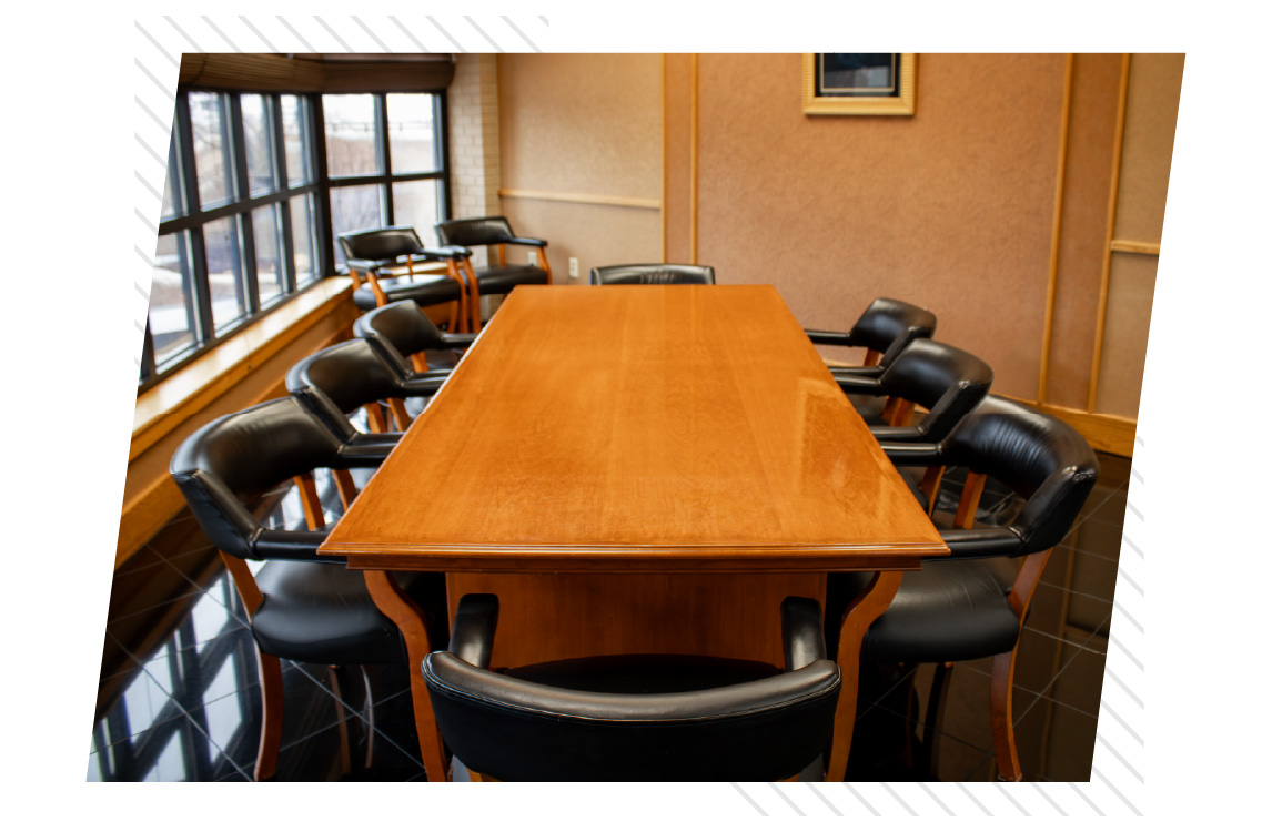 Rectangular wooden conference table surrounded by black leather chairs in a small meeting room with large windows along one wall and framed artwork on a tan wall.