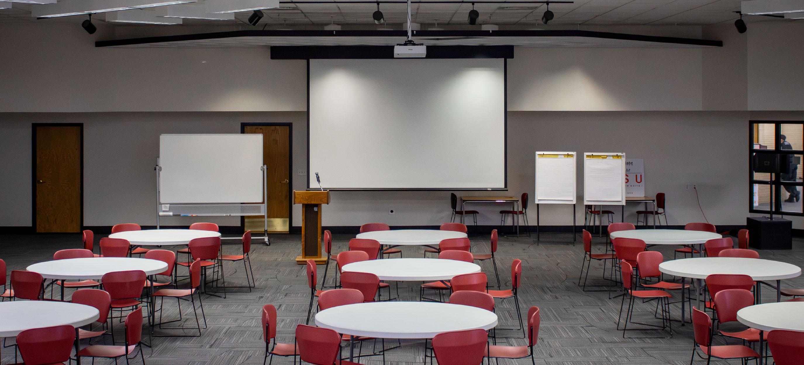 Large multipurpose event room set up banquet-style with round tables and red chairs, facing a projection screen, podium, whiteboard, and flip charts at the front.