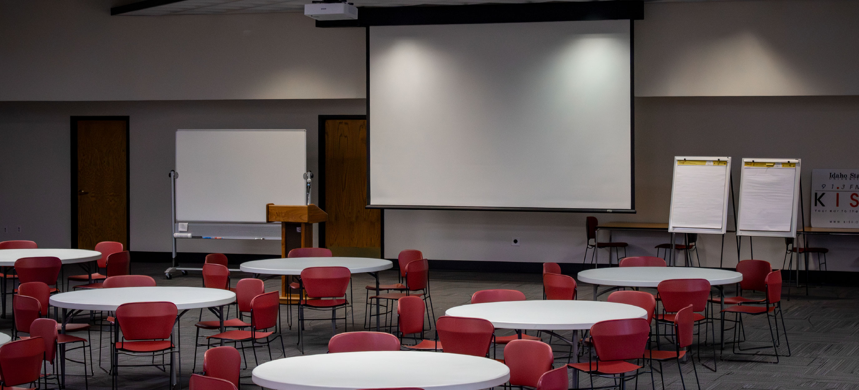Event room arranged banquet-style with round tables and red chairs, facing a large projection screen, podium, whiteboard, and two flip charts at the front.