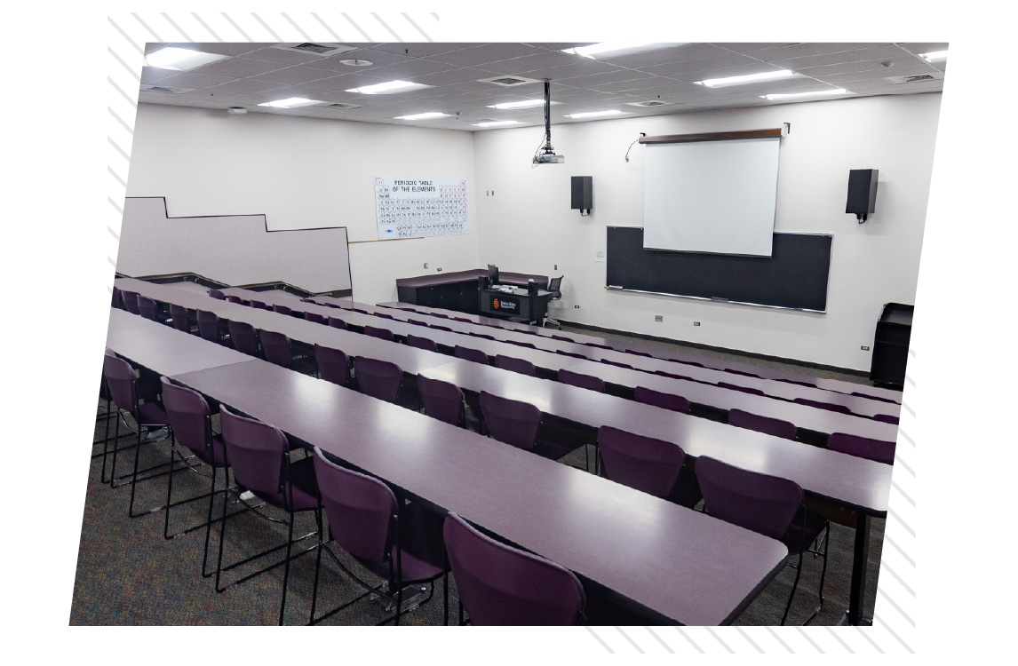Tiered classroom with long rows of tables and purple chairs arranged stadium-style, facing a projection screen, chalkboard, and instructor podium at the front of the room.
