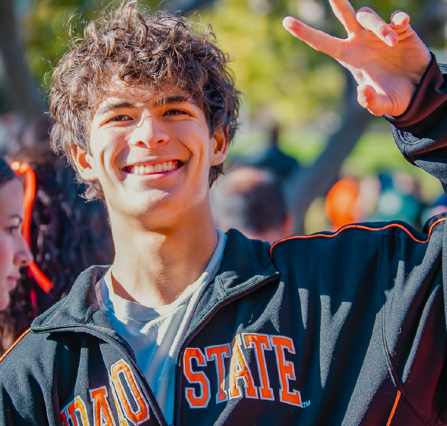 Smiling Idaho State University student wearing a black ‘Idaho State’ jacket makes a peace sign while standing outdoors on campus with other students in the background.