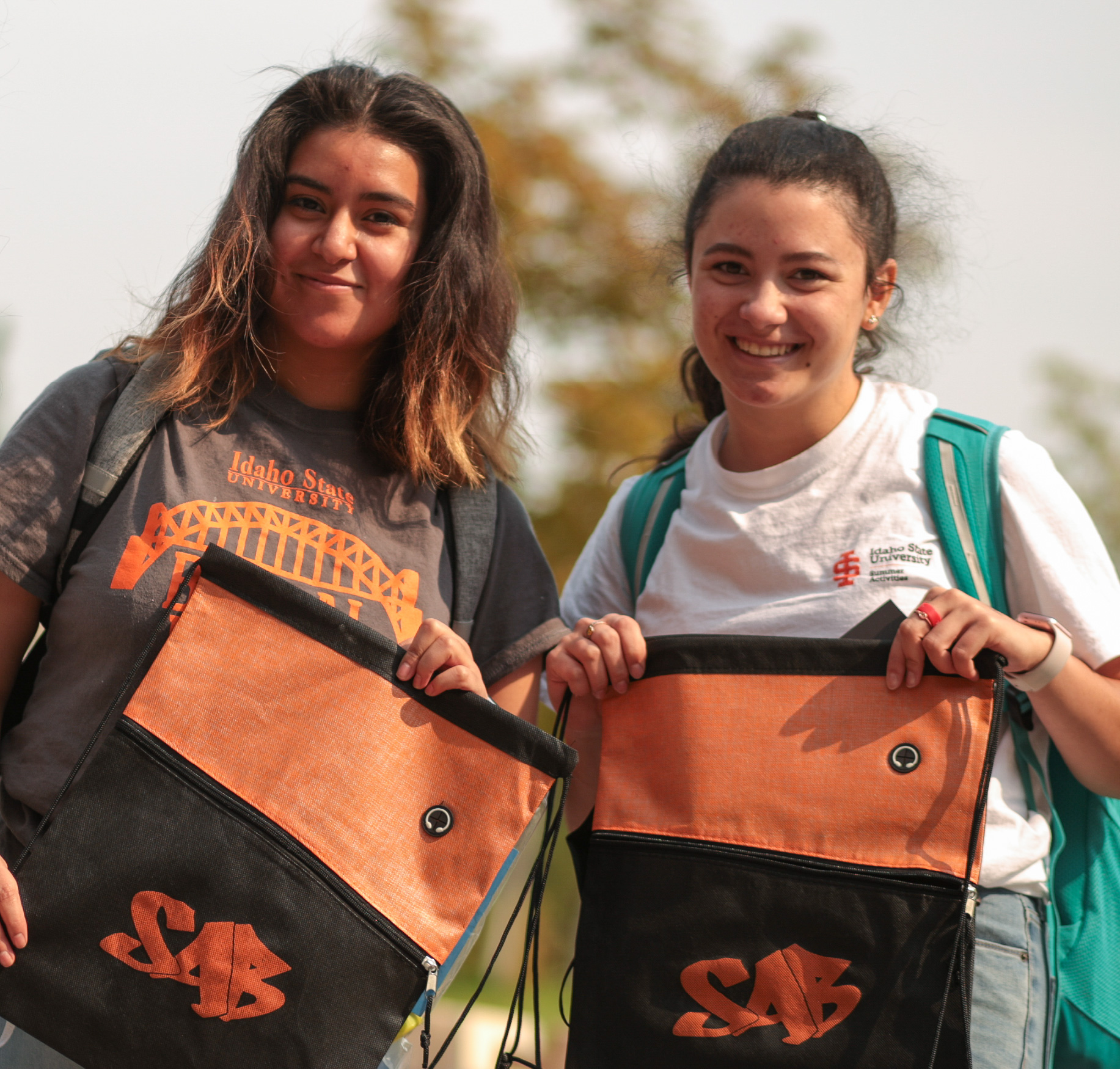 Two college students stand outdoors on campus smiling and holding orange and black SAB drawstring bags. Both wear backpacks, with trees softly blurred in the background, highlighting student involvement and campus activities