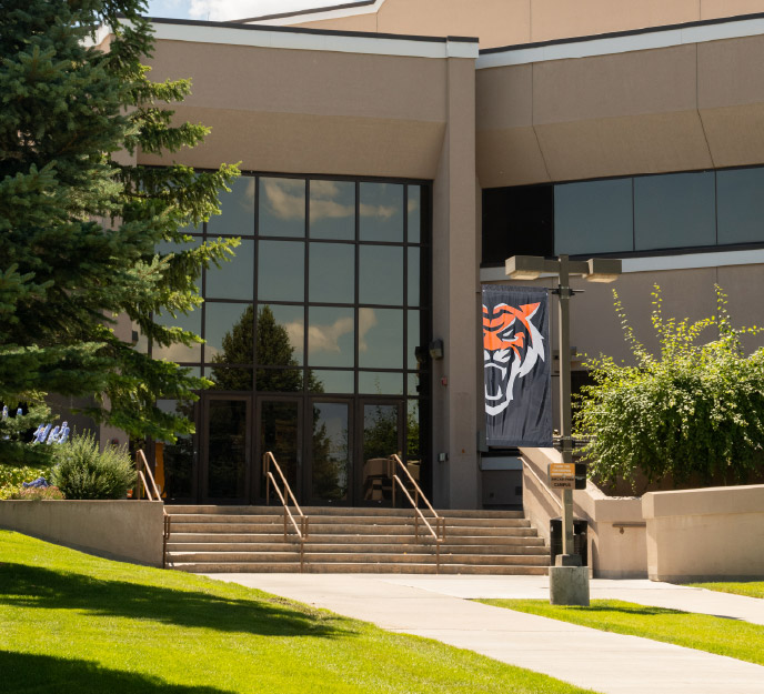 Front entrance of the Center for Higher Education (CHE) building at Idaho State University Idaho Falls, featuring a glass facade, wide stairway with handrails, landscaped greenery, and a Bengal tiger banner near the doorway.