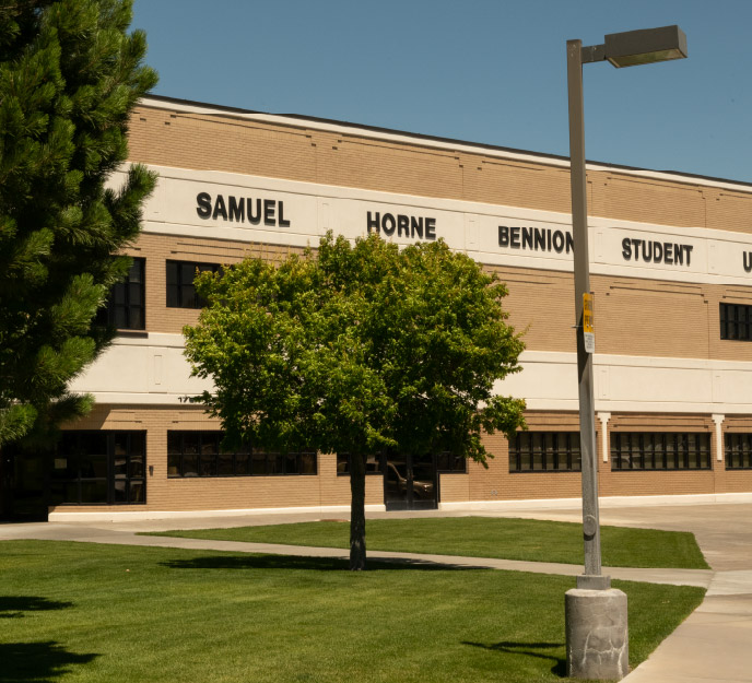 Exterior of the Bennion Student Union Building (BSUB) at Idaho State University Idaho Falls, showing the building name across the upper facade, a grassy lawn and walkway in the foreground, and trees and a campus light post near the entrance.