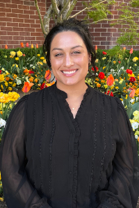 A woman with dark hair pulled back into an up-do and brown eyes smiles brightly in a portrait outdoors. She is wearing a black long-sleeved blouse with ruffled trim around the V-neck and delicate lace detailing on the front, along with a small gold necklace. Behind her is a vibrant garden bed filled with colorful red, orange, and yellow tulips and pansies, set in front of a red brick building wal