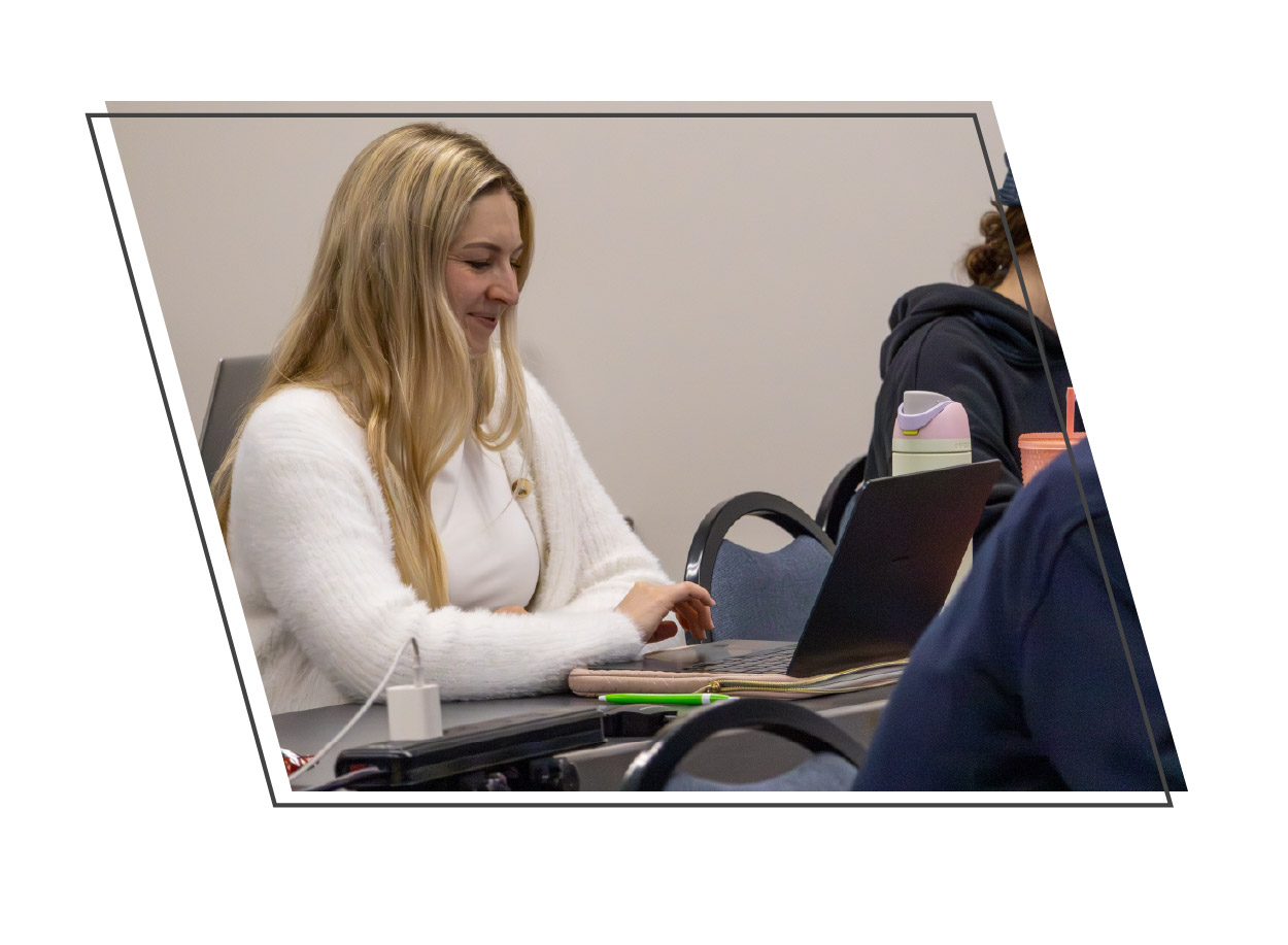 Student in a white sweater smiling while working on a laptop at a classroom table, with other students seated nearby. The photo is framed by an angled geometric border on a white background.